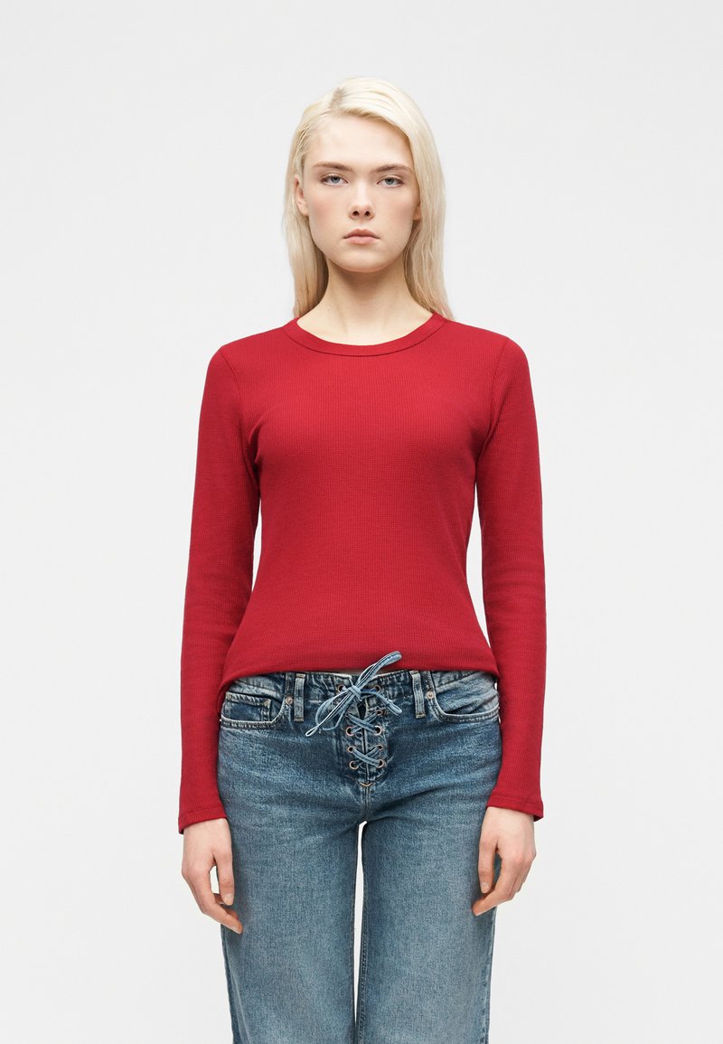 Ξανθιά γυναίκα που στέκεται μπροστά, φοράει εφαρμοστό κόκκινο πουκάμισο με μακριά μανίκια και ψηλόμεσα μπλε τζιν με δέσιμο μπροστά.