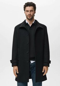 Abrigo de lana negro con un cuello alto, botones en la parte frontal y bolsillos laterales. Presenta una textura suave y un corte ajustado, superpuesto a un suéter oscuro.