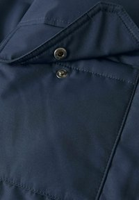 Tejido azul marino con una textura suave que presenta un cierre a presión y un bolsillo en el pecho. Se puede ver un detallado bordado a lo largo de los bordes.