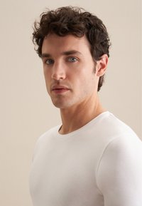 T-shirt blanc à manches longues avec un col rond, design ajusté, texture lisse et légère élasticité ; porté par une personne ayant les cheveux courts et bouclés.