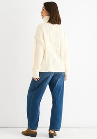 Chunky cable knit sweater in cream, featuring a high turtleneck and textured patterns. Paired with loose-fitting blue jeans and brown heeled mules.