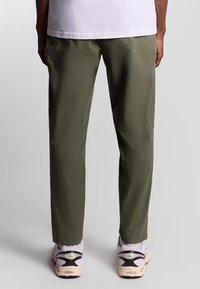 Olijfgroene broek met een gladde textuur, slim fit en enkellang, gecombineerd met grijze en crèmekleurige sneakers met zwarte accenten.