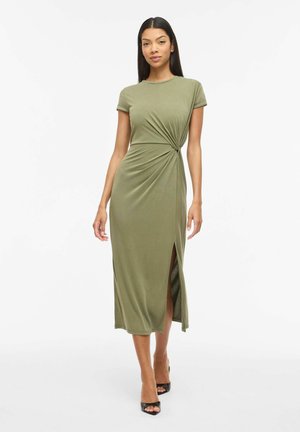 Naine, kes kannab oliivrohelist lühikeste varrukatega midi-kleiti, millel on külje lõhik ja vöökoht kokku tõmmatud detail, paistab koos mustade avatud varvastega kontsadega.