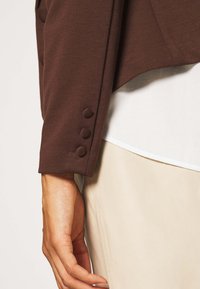 Brun blazerkant med tre rundade knappar klädda i tyg. Kanten har en slät yta, i kombination med ett ljust plagg under.