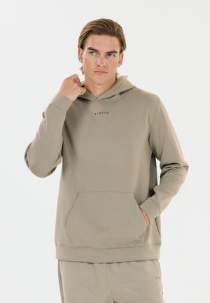 Young man wearing beige hoodie and matching pants with "Virtus" logo, standing with one hand in pocket and the other touching the hood.