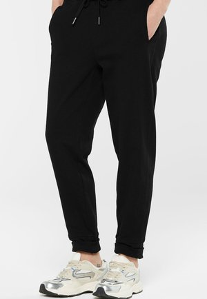 Person wearing black jogger pants with hands in pockets and white sneakers with silver accents, standing against a white background.