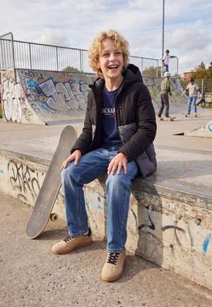 Un garçon souriant aux cheveux bouclés est assis à côté d’un skateboard dans un skatepark couvert de graffitis, tandis que d’autres jeunes font du skate à l’arrière-plan.