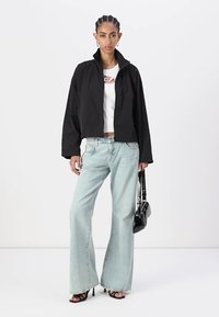 Giacca nera corta in materiale leggero, abbinata a una maglietta bianca e jeans a campana leggeri di colore blu chiaro. Accessoriata con una borsa nera.