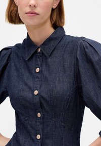 Rochie din denim cu corset strâns pe corp, mâneci scurte bufante și guler clasic. Are șapte nasturi maro pe partea din față și cusături verticale.