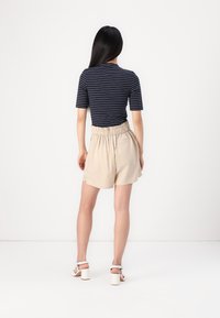 Navy striped fitted shirt paired with beige elastic-waist shorts. Model wears white block-heeled sandals. Minimalist design, smooth fabric.