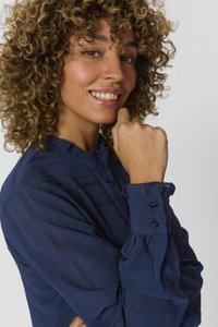 Blusa blu navy con righe texturate, colletto arricciato e polsini con bottoni. Il tessuto è leggero e leggermente trasparente, offrendo un aspetto sofisticato.