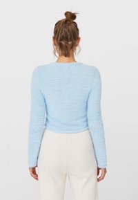 Suéter azul claro y esponjoso con mangas largas y escote redondo, que presenta una textura suave y un corte cropped, combinado con pantalones blancos.
