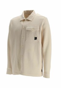Chemise beige à manches longues texturée avec boutons à l'avant, une poche poitrine boutonnée et un petit patch logo noir sur la poche.