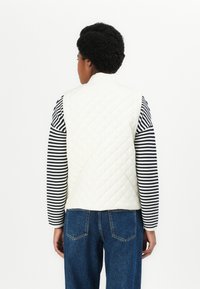 Gequilted crèmekleurige vest met een hoge kraag, gedragen over een zwart-wit gestreepte langekoop shirt. Blauwe jeans zichtbaar aan de onderkant.