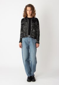 Svart kofta med vita geometriska mönster, krage, knäppning framtill, i kombination med ljusblå baggy jeans och svarta plattformsskor.