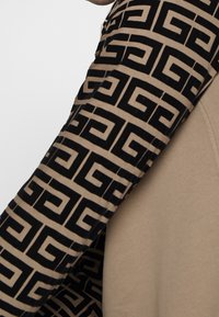 Maglia a maniche lunghe con corpo beige e tessuto nero a fantasia sulle maniche, con un design geometrico che attraversa tutta la fantasia.