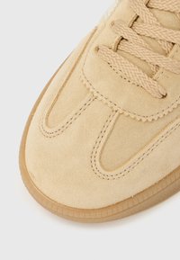 Nærbillede af beige ruskindsneakerspids med detaljeret syning, flettede snørebånd og en gennemsigtig gummisål.