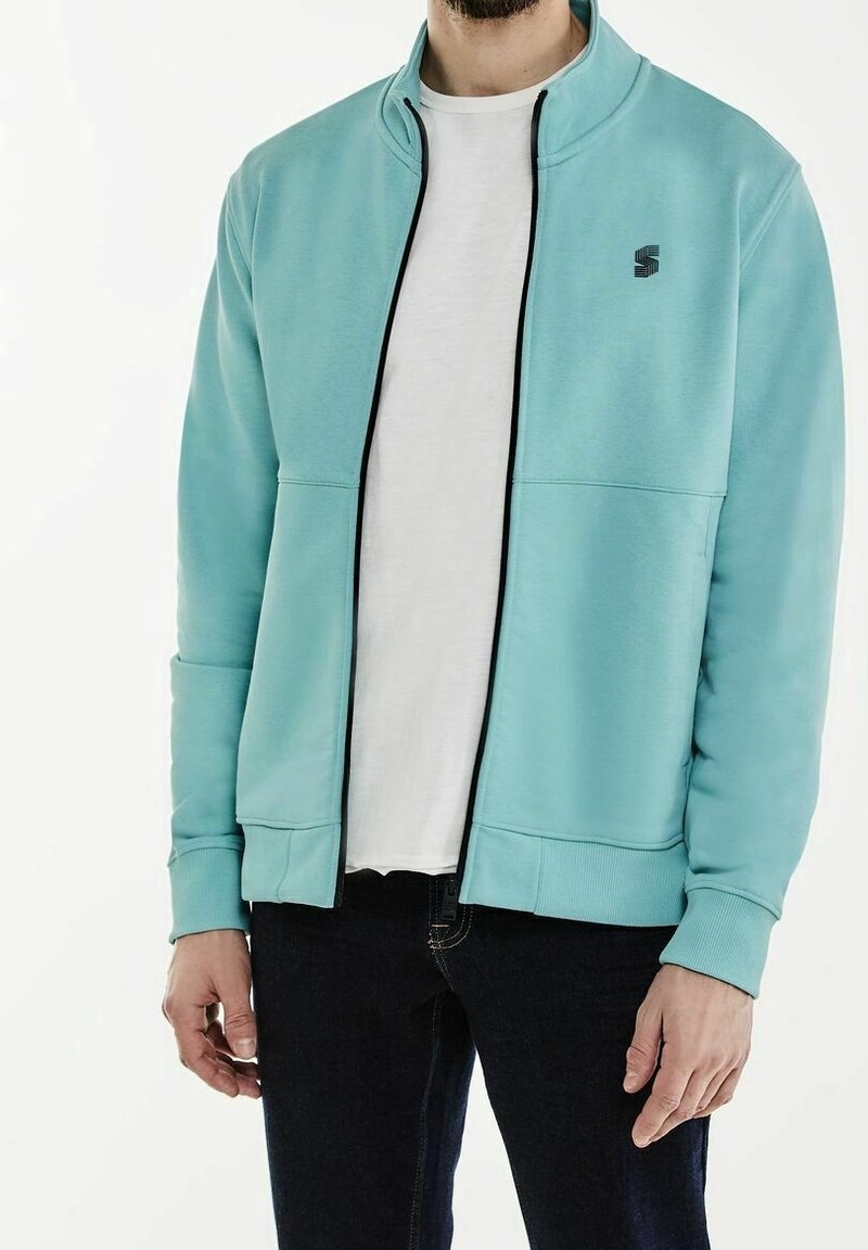 Light blue zip-up jacket in a smooth fabric, featuring a high collar, black zipper, and logo on the upper left chest. Ribbed cuffs and hem.