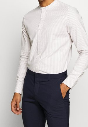 Uomo che indossa una camicia beige chiaro a righe con colletto alla coreana infilata in pantaloni eleganti blu scuro, con una mano in tasca.