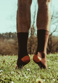 Brūnas kokvilnas zeķes ar oranžiem akcentiem uz papēžiem, parādītas uz kājām, stāvot uz zālāja ar maziem baltajiem ziediem.