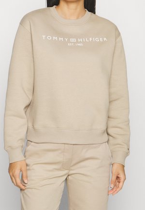 Persoon draagt een beige Tommy Hilfiger-trui met witte logotekst en bijpassende beige broek, staand tegen een effen achtergrond.