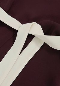 Burgundin kangas sileällä tekstuurilla, jossa on valkea vyö, taitavasti solmittuna keskelle, luoden voimakkaan kontrastin.