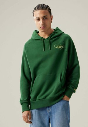 Sudadera verde con bolsillo frontal, capucha ajustable y logo "Kappa" en dorado. Fabricada en material suave con puños y dobladillo acanalados. Llevada con jeans azules.