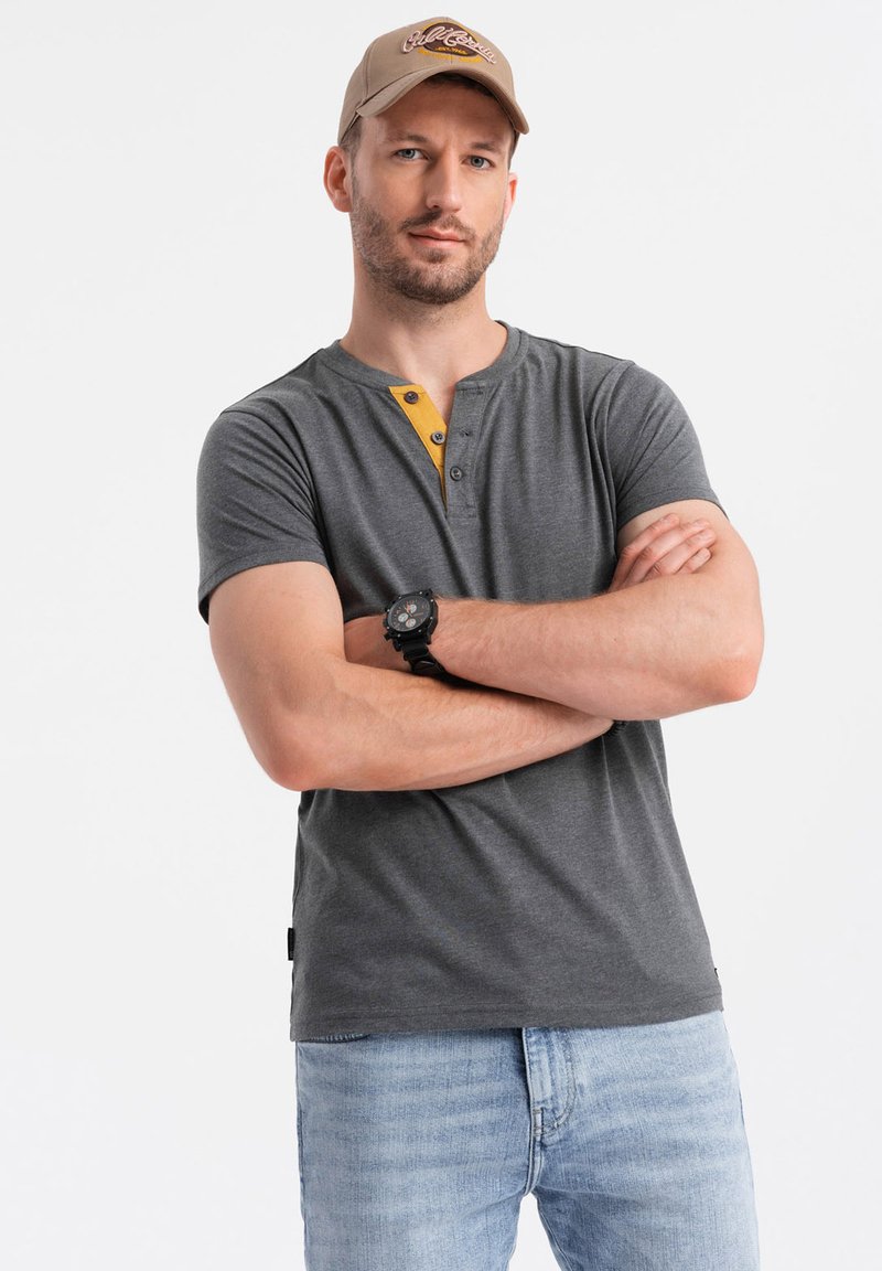 Camisa henley gris de manga corta con una tapeta de botones amarillos, combinada con jeans azul claro. El modelo lleva una gorra beige y un reloj negro.