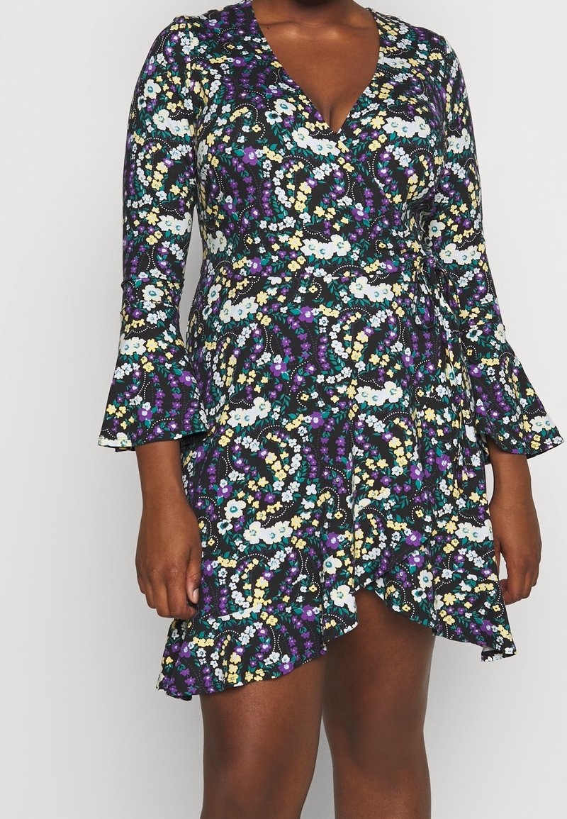 Femme portant une robe portefeuille à manches longues, avec des fleurs violettes, blanches, jaunes et bleu canard, et un ourlet à volants.