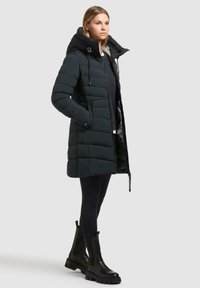 Giubbotto modesto nero con colletto alto, caratterizzato da una chiusura a zip, tasche laterali e un motivo trapuntato. Indossato con leggings neri aderenti e ankle boots neri spessi.