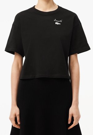 Kobieta nosząca czarny krótki T-shirt z małym białym logo Lacoste na lewej piersi oraz czarną spódnicę.