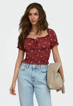 Jeune femme aux longs cheveux ondulés portant un haut smocké à fleurs bordeaux, un jean taille haute bleu clair, et tenant un sac beige plié.