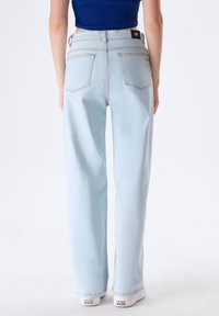 Jeans de tiro alto y pernera ancha en un azul claro, con bolsillos traseros de parche, detalles de costura sutiles y una etiqueta negra en la cintura. Combinados con zapatillas blancas.