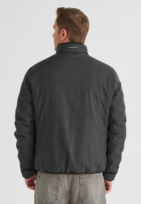 Schwarze isolierte Jacke mit hohem Kragen, mit einer gesteppten Textur und Logo auf dem Rücken, kombiniert mit hellgrauen Jeans.