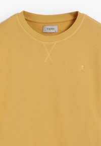 Sudadera amarilla con cuello redondo acanalado, textura de punto ligera y un pequeño logo bordado de calavera y huesos cruzados en el pecho.