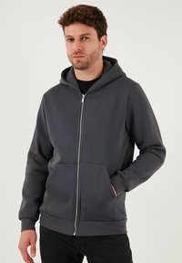 Sivi zip-up dukserica od glatkog materijala, sa zatvaračem sprijeda, dva prednja džepa i kapuljačom svezanom špagom. Tekstuirani manžeti i rub.