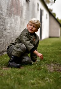 Dětská prošívaná bunda olivově zelené barvy, doplněná o kalhoty ve stejném odstínu a černé gumové boty. Dítě je v dřepu na trávě a sahá po nějakém předmětu.