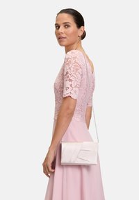 Top in pizzo rosa chiaro con maniche corte abbinato a un lungo vestito coordinato. La modella tiene in mano una piccola borsa elegante in satin color crema.