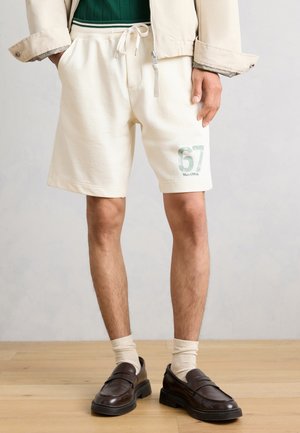 Mand iført cremefarvede shorts med "67 Marc O'Polo" print, beige jakke, mørkebrune loafers og cremefarvede sokker, der står på trægulv.