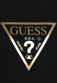 Sudadera negra con un gran logo triangular dorado que muestra "GUESS", "U.S.A.", y "WASHED JEANS" en texto blanco y holográfico, diseño centrado.