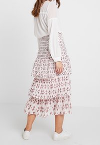 Ker skirt berlapis bertingkat berwarna merah muda pucat dengan pola geometris merah. Kain ringan dengan desain mengalir, dipadukan dengan blus putih lengan panjang.