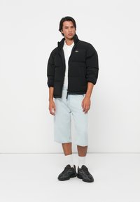 Veste rembourrée noire avec un col haut, fermeture éclair à l'avant et logo vert, associée à un short en denim clair et des baskets noires.