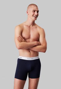 Boxeri bleu marin cu un brâu elastic alb pe care este inscripționat logo-ul „Calvin Klein”. Țesătura pare netedă și elastică. Modelul stă cu brațele crossing.