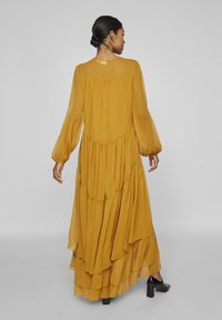 Vestido maxi amarelo em camadas com longas mangas transparentes. Apresenta tecido fluido e um decote alto, detalhado com suaves pregas na parte de trás.