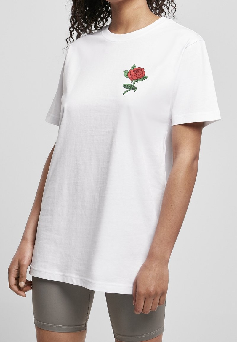Femme portant un t-shirt blanc uni avec un petit motif de rose rouge sur la poitrine et un short gris moulant sur un fond uni.