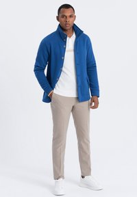Chaqueta de forro polar azul con cuello alto y cierre de botones, llevada sobre una camiseta blanca tipo polo. Unos pantalones beige y zapatillas blancas completan el atuendo.