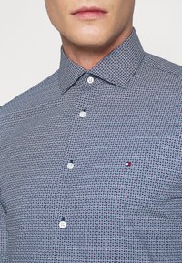 Laivastonsininen nappipaita, jossa on pieniä valkoisia ja punaisia kolmiokuvioita. Paidassa on leveä kaulus ja valkoiset napit.