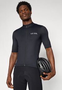 Maillot de cyclisme noir à manches courtes avec zip, arborant un logo blanc sur la poitrine gauche. Le cycliste tient un casque noir avec des aérations.