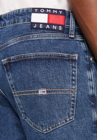 Jeans i denim med en dyp blå farge, med en baklomme og en rektangulær Tommy Jeans-label med røde, hvite og blå detaljer.