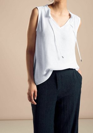 Kobieta w białej, bezrękawnikowej, teksturowanej bluzce z kapturem i sznurkami oraz czarnych, teksturowanych szerokich spodniach, z jedną ręką w kieszeni, na beżowym tle